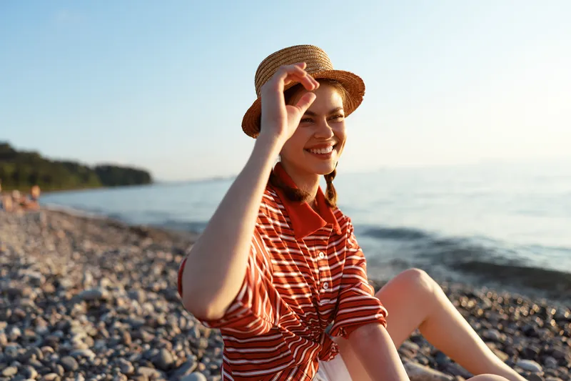 A smiling young woman in a straw hat sits on a pebble beach by the sea. Lifestyle concept of summer vacation, outdoor travel, relaxation and happiness in warm sunlight.