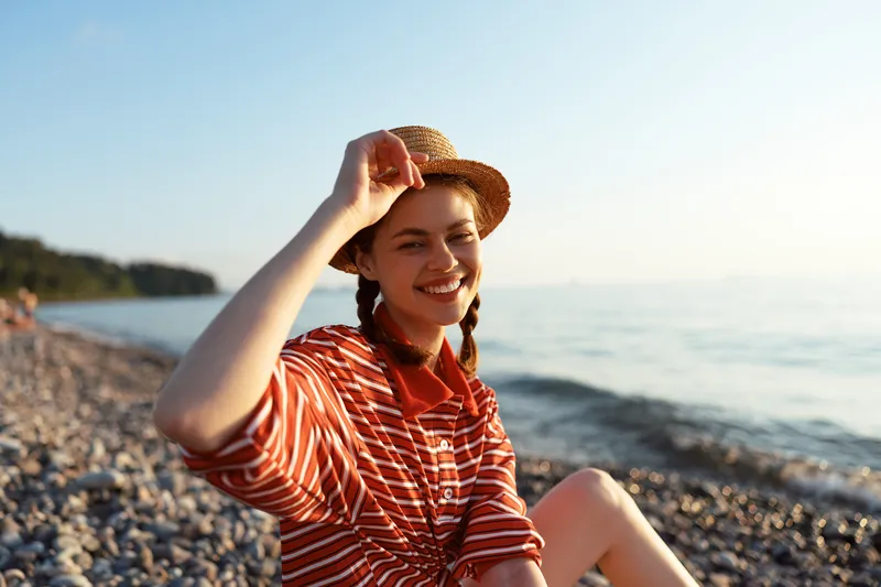 A smiling young woman lifestyle portrait wearing a straw hat on a pebble beach at sunset. Concept of summer vacation, travel, freedom, joy and leisure near ocean.
