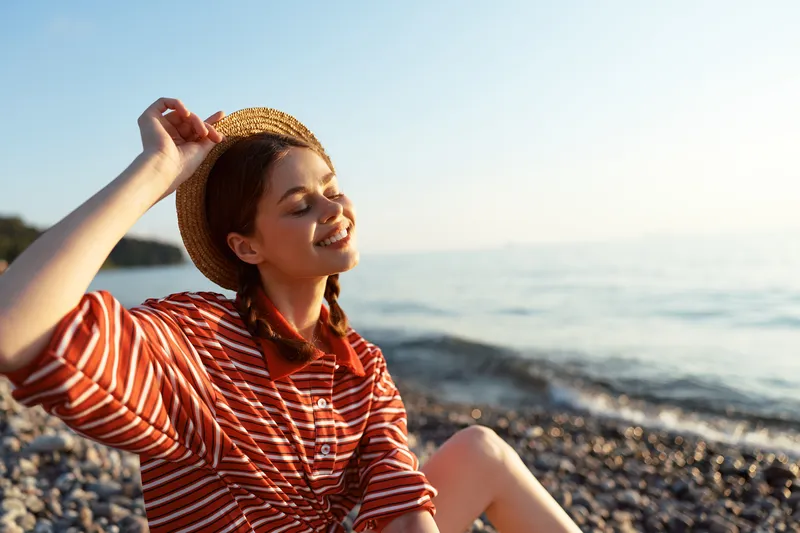 Young woman in a striped shirt and straw hat relaxes on a sunny pebble beach by the sea, enjoying the warm light and peaceful atmosphere for a summer lifestyle.