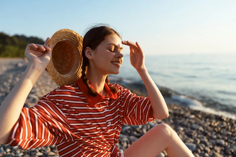 Smiling woman enjoying lifestyle on pebble beach at sunset. She holds straw hat, wearing red striped shirt. Summer vacation concept, warm sunlight and peaceful atmosphere near sea, soft lighting.