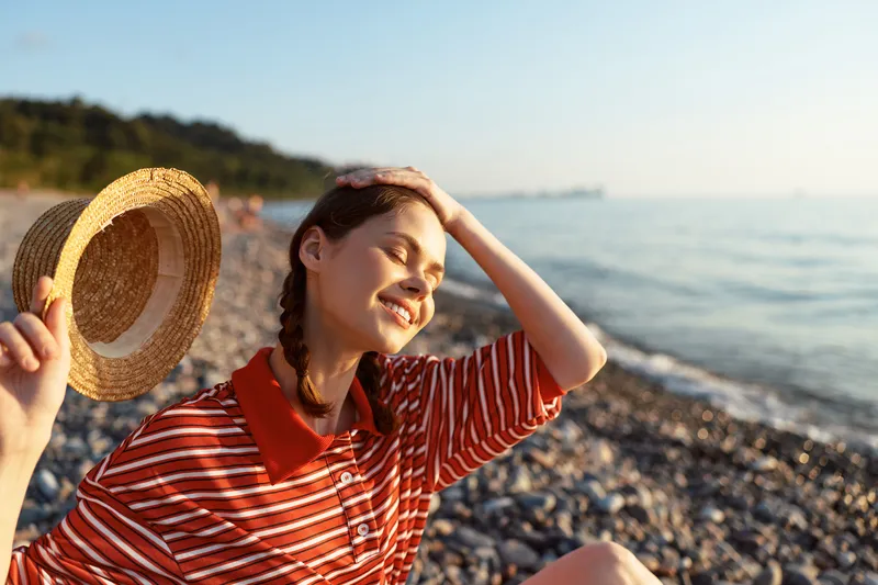 Smiling woman enjoying sunny summer beach, holding straw hat, wearing red striped shirt. A lifestyle portrait reflecting joy, relaxation, travel, happiness, and outdoor wellness.