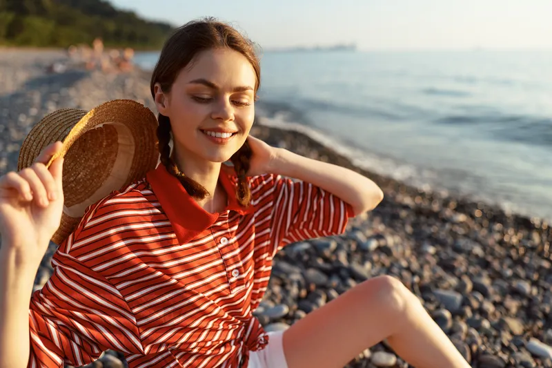 Smiling young woman in striped shirt wearing straw hat sitting on pebble beach. Happy lifestyle portrait at sunset. Concept of summer vacation, relaxation, nature, holidays and travel.