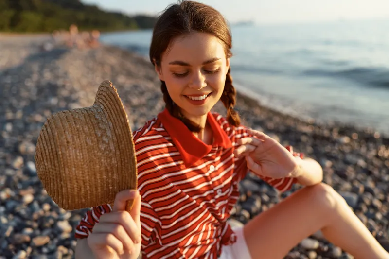 Young woman, lifestyle, portrait, beach, hat, smiling happily on a pebbly shore at sunset. Atmospheric outdoor lifestyle photo of a girl resting during summer vacation by the ocean sea.