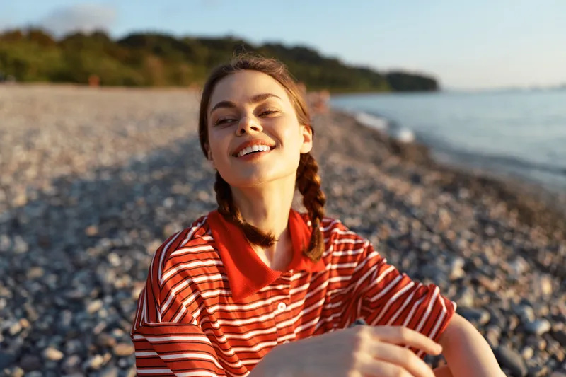 Smiling woman lifestyle portrait at the beach. Happy girl with braids sitting on pebbles near the ocean. Relaxed summer leisure, sunshine, vacation, travel concept.