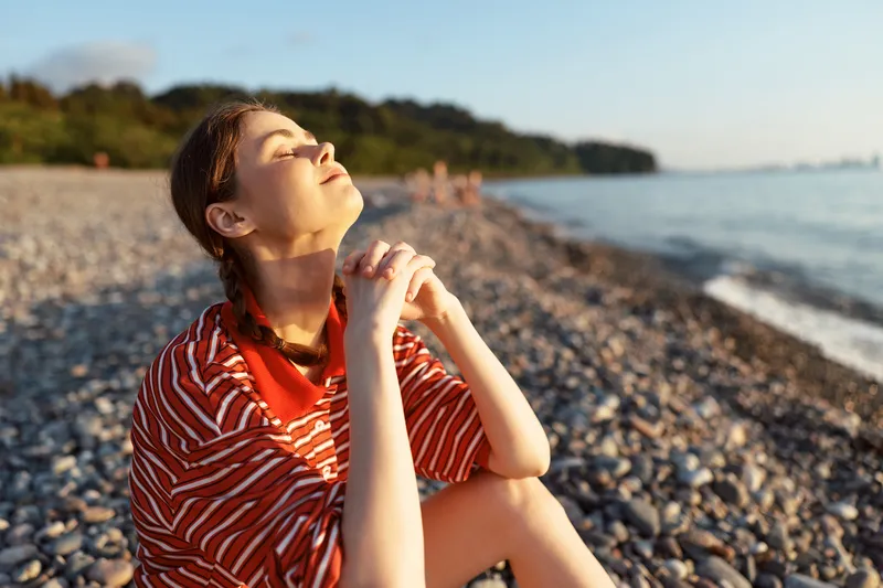 A young woman lifestyle portrait sitting on a pebble beach at sunset. She is calm, peaceful, eyes closed, relaxing outdoors near the sea. Concept for wellness, freedom, nature.