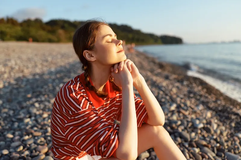Young woman enjoying lifestyle on rocky beach during sunset. She sits with closed eyes feeling peace and happiness. Warm soft light illuminates her face. Concept for travel or summer vacation.