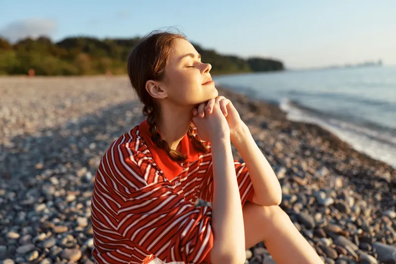 A happy young woman relaxing on a pebble beach at sunset. She enjoys the warmth of sunlight outdoors, lifestyle photography capturing peaceful moments by the calm sea water.