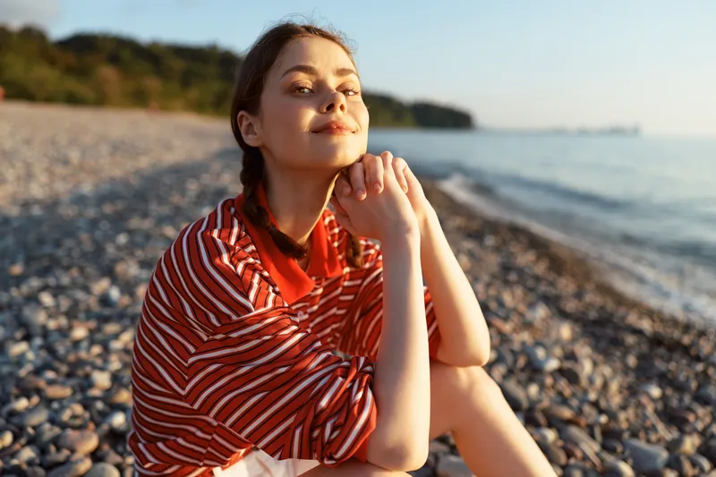 Young woman enjoying lifestyle on pebble beach at sunset. Relaxed lady sitting outdoors, resting, looking at camera. Golden hour glowing light. Concept of harmony and travel.