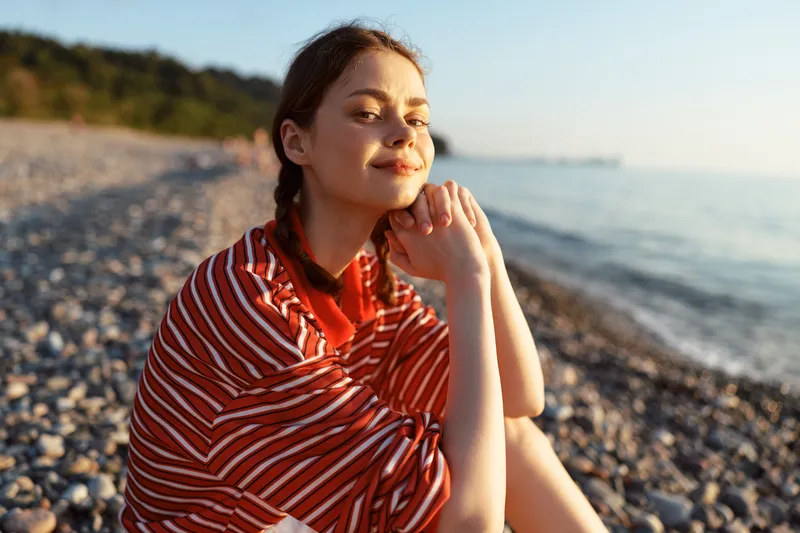 A smiling young woman lifestyle portrait on a pebble beach at sunset. She enjoys the golden hour sea breeze. Concept of vacation, freedom, relaxation, and summer beauty.
