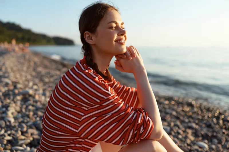 A smiling young woman lifestyle portrait sitting on a stony beach during a sunny golden sunset, looking blissful and relaxed. Concept for summer vacation, seaside travel, and peaceful joy.