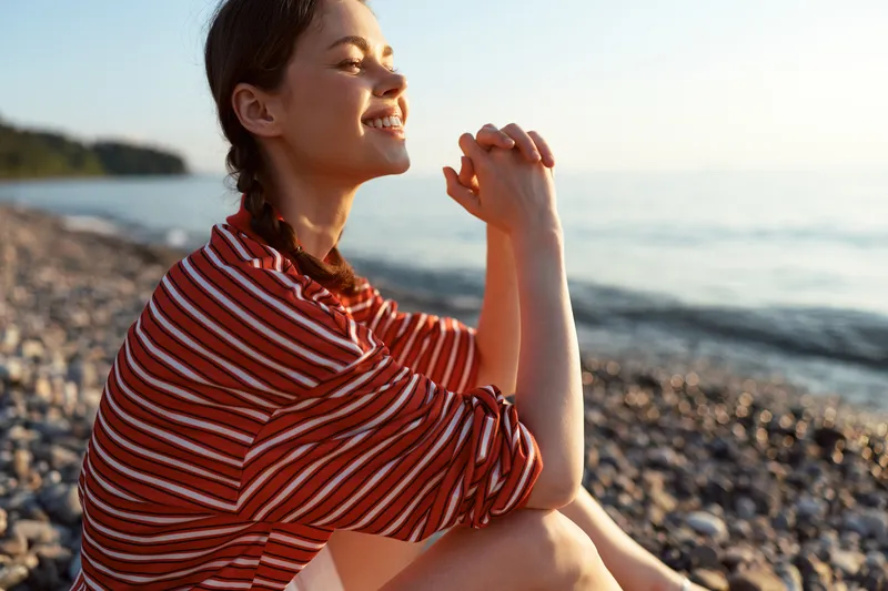 Young woman with braided hair relaxes on a pebble beach at sunset, smiling thoughtfully while sitting near the water. A peaceful lifestyle shot capturing a calm seaside moment.