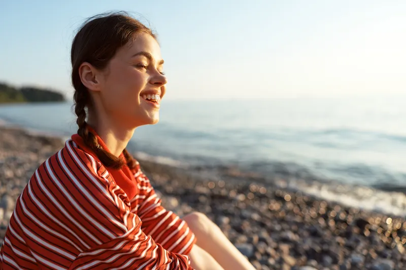 A happy woman relaxing on the pebble beach at sunset lifestyle concept, enjoying the peace and warm summer sunlight near the sea water, smiling and feeling calm in a beautiful golden hour.
