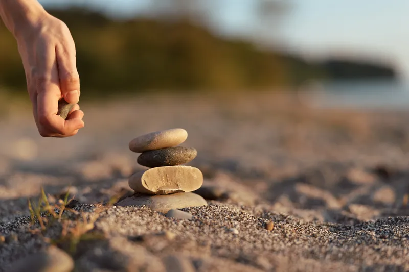 A woman lifestyle detail as she stacks stones on a beach. Balanced rocks placed on sand at sunset representing mindfulness, zen, meditation, calm, harmony, peace, and serenity.