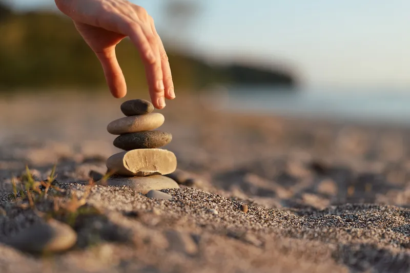 A woman builds a balanced stone stack on a beach, hand placing a pebble for stability. Relaxed lifestyle concept focusing on meditation, harmony, zen, nature, mindfulness, and balance.