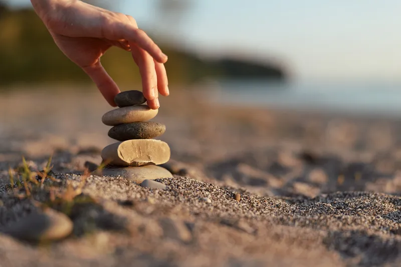 Woman lifestyle concept detail where a hand balances smooth zen stones on a sandy beach. Peaceful outdoor meditation scene at sunset light creating a calm and mindful atmosphere.