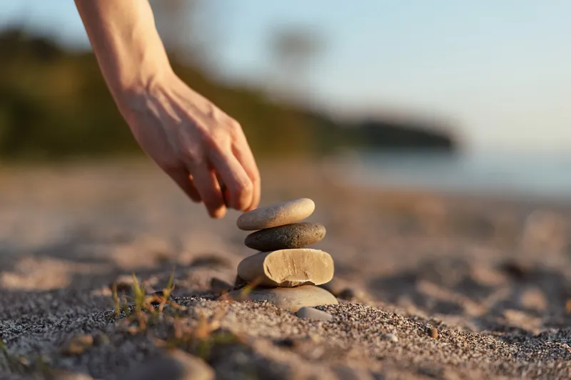 Woman hand stacking stones on beach for balance and zen. Lifestyle concept of meditation, peace, and harmony. Golden hour sunlight creates a calm and mindful atmosphere outdoors.