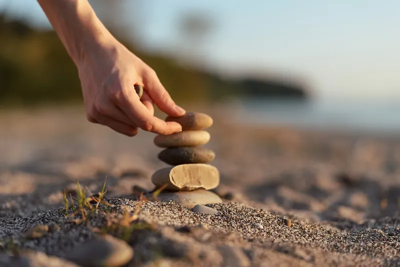 A young woman balances smooth stone stack on sunny beach shore. Female hand builds zen cairn during peaceful lifestyle moment for meditation, balance, and wellness.