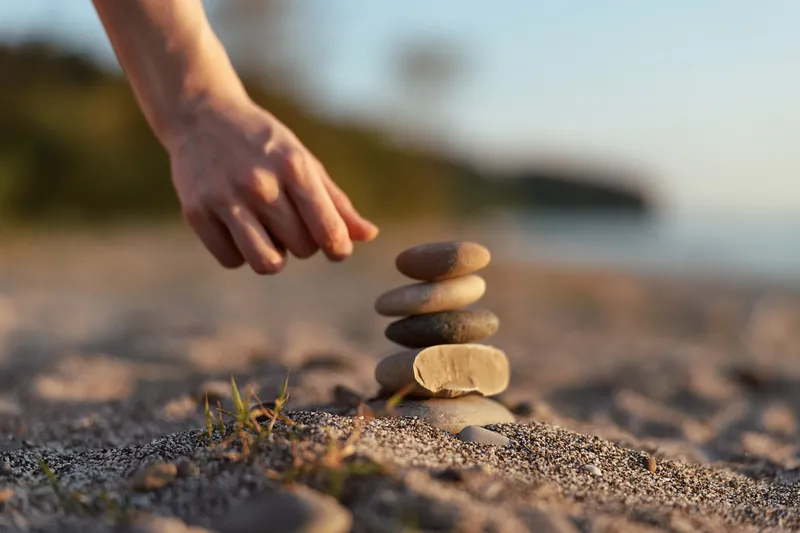Woman lifestyle concept showing hand stacking balancing stones on sandy beach. Tranquil scene with rock pile and blurred ocean background. Natural lighting creates peaceful Zen meditation mood.