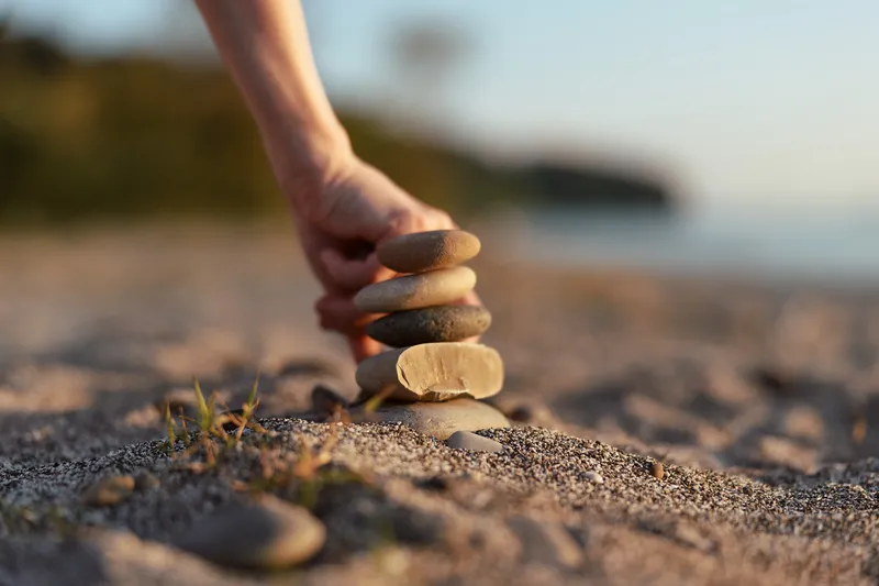 Woman lifestyle concept showing a hand stacking natural stones on the beach at sunset. The balancing zen rocks create a calm atmosphere during travel and relaxation on the sandy ocean shore.