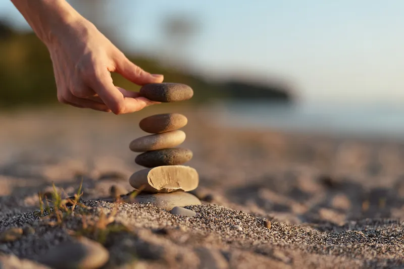 Woman stack stones on beach for balance and harmony, lifestyle tranquility concept. Hand places pebble on zen tower at sunset hour. Blur background with sand and warm light.