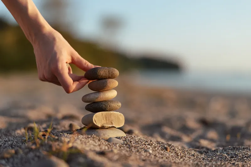 A woman lifestyle hand balancing smooth stones on a sandy beach. Conceptual image for zen meditation, spa, wellness, harmony, balance, patience, and focus during a calm sunset.