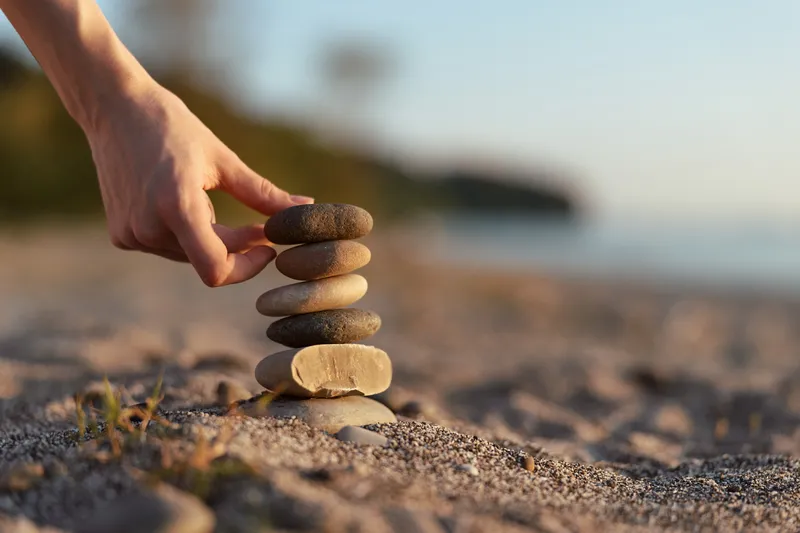 Woman building stone balance tower on beach at sunset. Lifestyle concept of zen, harmony, peace, and mindfulness with blurred ocean background. Soft natural golden lighting.