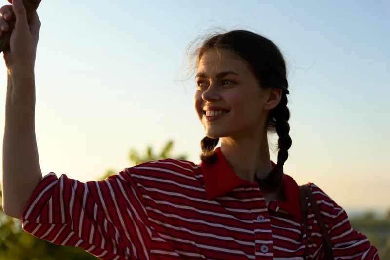Young woman with braided hair wearing a striped shirt smiles outdoors during golden hour. A happy portrait capturing her lifestyle while looking at the bright horizon in warm sunset light.