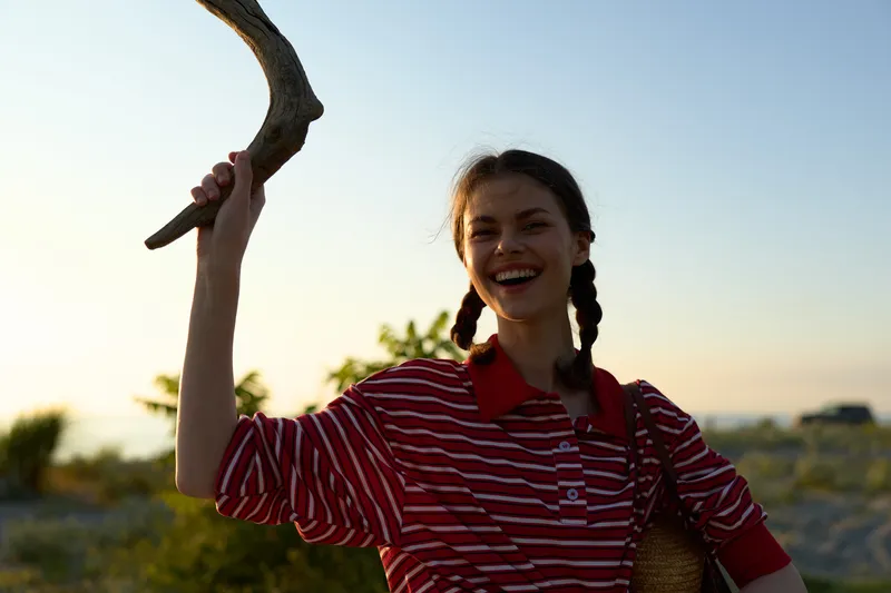 Happy woman lifestyle portrait holding wooden branch outdoors in summer. The joyful lady with braided hair smiles under sunset soft golden light on a beach landscape.
