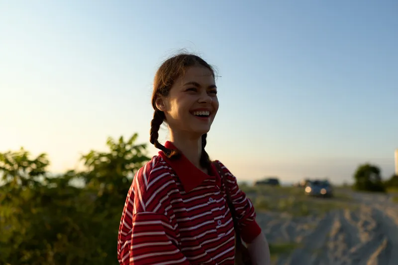Cheerful young woman with braided hair wearing a striped polo shirt smiles outdoors during sunset light. Concept for authentic lifestyle and happy summer vacation moments.