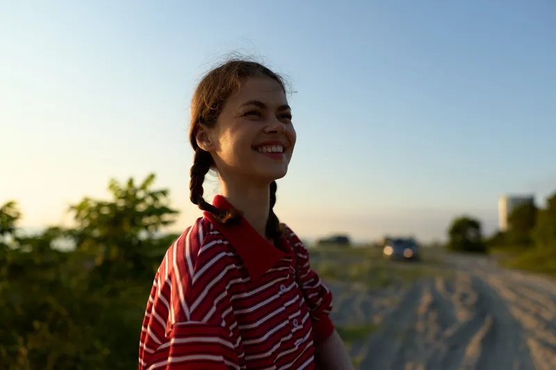 Smiling woman lifestyle portrait outdoor at sunset. Young female with braids enjoys summer vacation in casual clothes. Natural golden hour light creates a happy, relaxed, authentic warm mood.