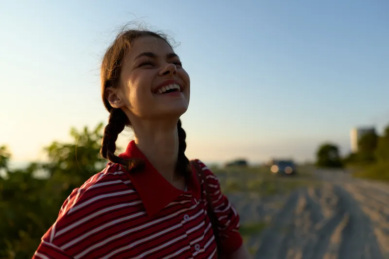 A joyful young woman with braided hair wearing a striped polo shirt laughs outdoors during sunset. The authentic lifestyle portrait captures her genuine happiness in a warm natural light.