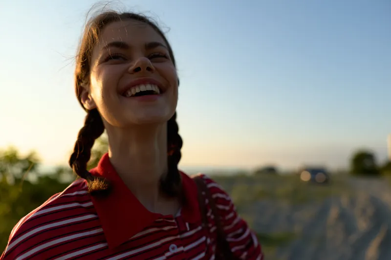A joyful young woman with braided hair laughs under golden sunset light, wearing a striped polo shirt while enjoying a peaceful moment in nature, reflecting authentic positive life energy.