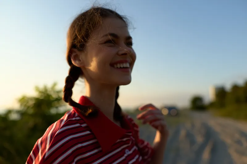 Happy woman enjoying lifestyle, smiling outdoors at sunset. Positive girl wearing striped shirt outdoors, braid hairstyle, natural lighting, summer concept for freedom, joy and summer mood.