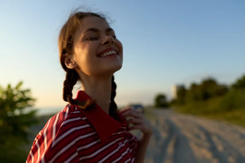 A happy young woman with braids enjoying sunset outdoors. Lifestyle portrait of a smiling female expresses freedom, joy, and peace in a natural summer environment.