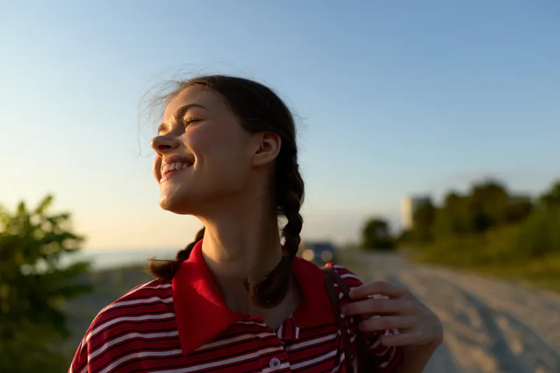 Happy woman lifestyle portrait outdoor at sunset. Young girl smiling with braids enjoys warm golden hour light on coastal road. Authentic candid feeling for travel or summer concept.