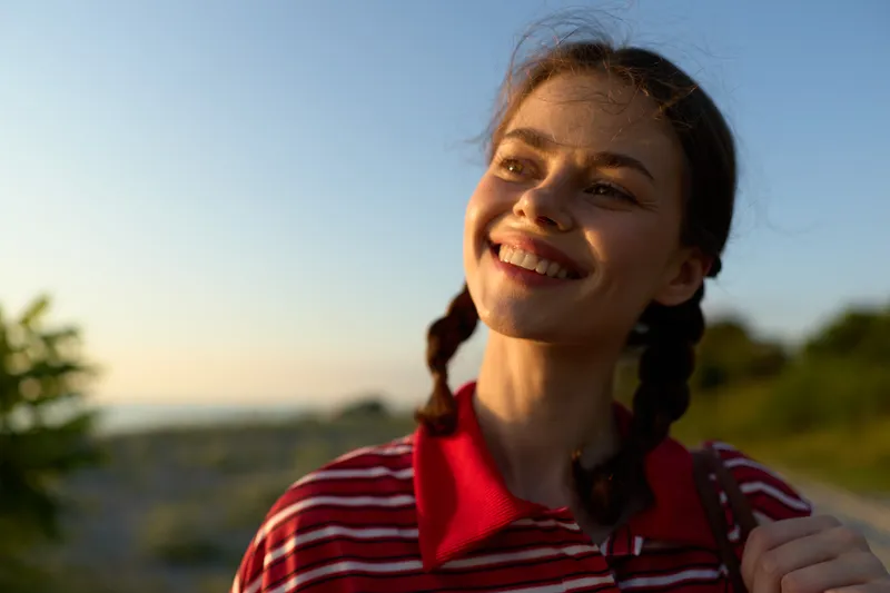 Young woman, lifestyle, portrait, sunlit, relaxing outdoors during sunset. The smiling girl looks away with a joyful expression. Concept for freedom, happiness, and peace.