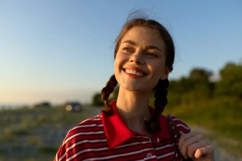 A smiling young woman enjoying lifestyle outdoors on a summer day. The happy female with braided hair looks at the sunlight, urban travel concept, authentic natural beauty portraits.