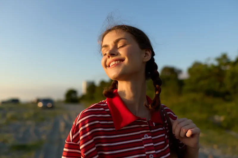 Smiling young woman enjoying summer evening sunset. Lifestyle portrait of happy girl outdoor in nature. Concept of freedom, peace, vacation, happiness and wellness.