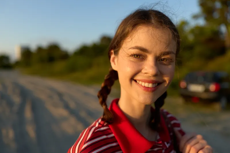 Smiling woman with braids posing outdoors during sunset. She enjoys lifestyle, nature, freedom, summer, happiness. Golden hour light creates a bright mood in this portrait photograph.