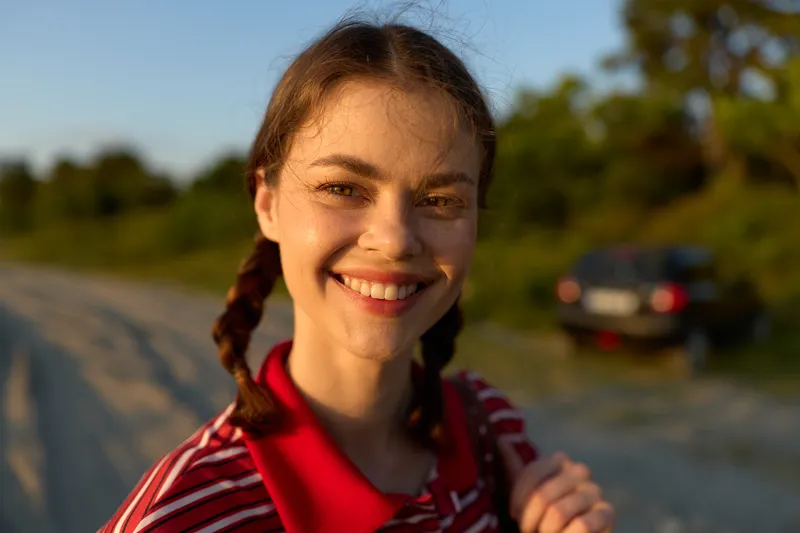 Smiling young woman with pigtails posing outdoors during sunset. Lifestyle photography capturing happy emotions and peaceful mood in warm golden evening light on a countryside road background.