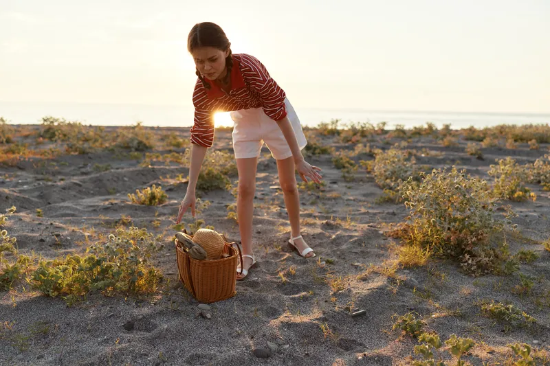 Young woman, lifestyle, picnic, basket, beach, sunset, standing on sandy ground, puts her hand near woven bag. Warm golden light, scenic dusk background, vacation, leisure, outdoors.