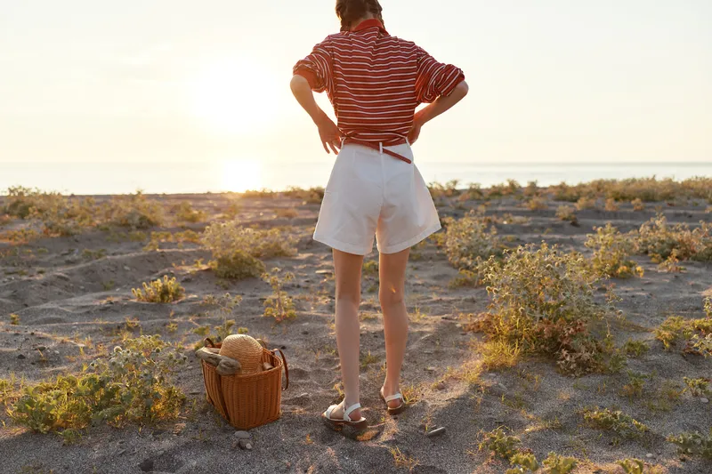 A young woman enjoying lifestyle on the sunny beach at sunset, standing near basket with bag. Concept for summer vacation, seaside recreation, freedom, travel, relaxation, and atmospheric day.