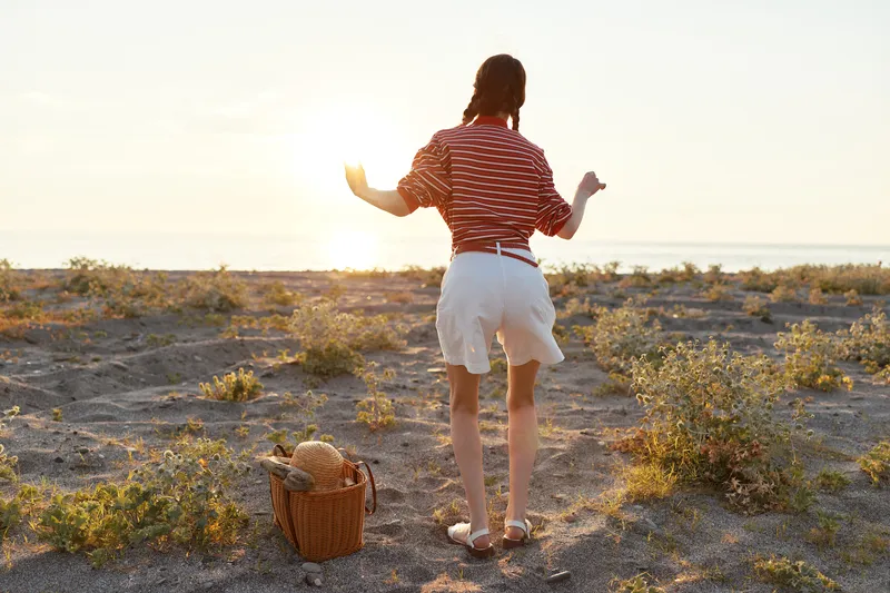 Young woman with braids wearing a striped shirt dances gracefully on a sandy beach at golden sunset hour while a picnic basket rests nearby in the peaceful coastal landscape.
